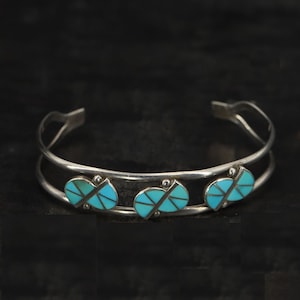 May include: A silver cuff bracelet featuring three turquoise leaf-shaped inlays. The bracelet has a double-band design and is set against a dark background. The turquoise inlays are a bright blue colour.