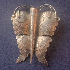 May include: A silver-toned butterfly brooch with detailed dot and line patterns. The wings are segmented and have a scalloped edge. The antennae are curved, and the body is a textured cylinder. The brooch is likely made of metal.