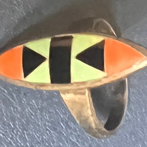 Pode incluir: Um anel prateado com um design em forma de olho. O olho apresenta formas geométricas laranja, pretas e verde claro. A banda do anel é simples e lisa.
