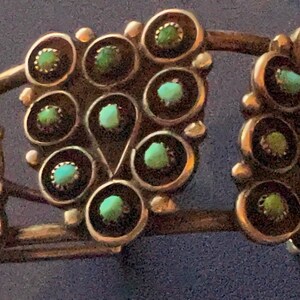 Könnte beinhalten: Silberfarbenes Armreif-Armband mit Türkissteinen. Das Armband hat eine Reihe von kreisförmigen Designs, jedes mit einem Türkisstein in der Mitte. Das Design wiederholt sich über das Armband und erzeugt ein symmetrisches Muster. Das Armband ist ein Schmuckstück.