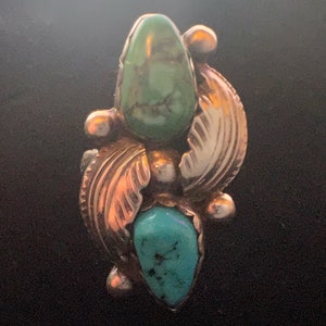 May include: A silver-toned ring featuring two turquoise-colored stones and two green-colored stones. The ring has a leaf-like design with silver-colored accents. The ring is set against a dark background.