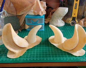 Puede incluir: Dos pares de cuernos de plástico beige moldeados, cada uno con una punta puntiaguda y una base curva. Los cuernos están dispuestos en un patrón simétrico sobre una estera de corte verde.