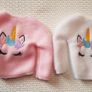 May include: Two doll-sized sweaters with unicorn embroidery. One sweater is pink and the other is white.