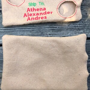 May include: Beige felt envelope-shaped pouch with embroidered details. The top portion features red and green text reading "Ship To: Athena Alexander Andres" and a circular stamp with "DEC" inside. Two circular cutouts are outlined in red.