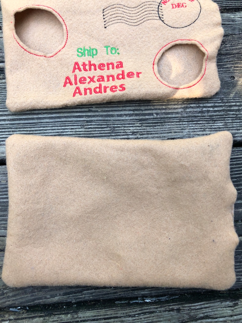 May include: Beige felt envelope-shaped pouch with embroidered details. The top portion features red and green text reading "Ship To: Athena Alexander Andres" and a circular stamp with "DEC" inside. Two circular cutouts are outlined in red.