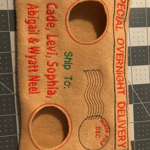 May include: Tan felt envelope with red and green embroidered text. The text reads "SPECIAL OVERNIGHT DELIVERY" and "Ship To: Cade, Levi, Sophia, Abigail & Wyatt Neel." Features three circular cutouts with red stitching.