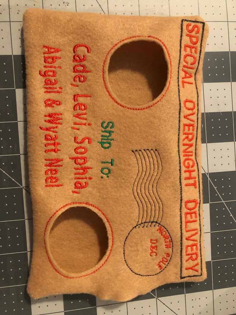 May include: Tan felt envelope with red and green embroidered text. The text reads "SPECIAL OVERNIGHT DELIVERY" and "Ship To: Cade, Levi, Sophia, Abigail & Wyatt Neel." Features three circular cutouts with red stitching.