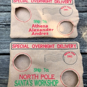May include: Two beige felt Christmas envelope-shaped pouches with red and green embroidered text. The top pouch reads "SPECIAL OVERNIGHT DELIVERY" and "Ship To: Athena Alexander Andres." The bottom pouch reads "Ship To: NORTH POLE SANTA'S WORKSHOP."