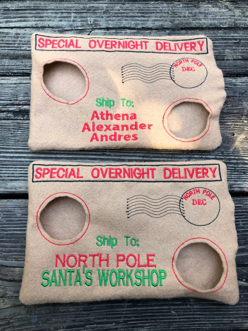 May include: Two beige felt Christmas envelope-shaped pouches with red and green embroidered text. The top pouch reads "SPECIAL OVERNIGHT DELIVERY" and "Ship To: Athena Alexander Andres." The bottom pouch reads "Ship To: NORTH POLE SANTA'S WORKSHOP."