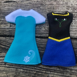 May include: Two felt doll dresses, one blue with a snowflake and swirl design, the other black with a yellow trim and green embroidery.