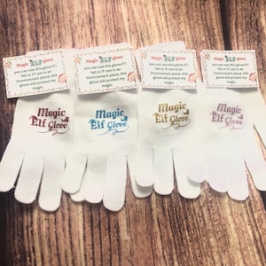 May include: Four white knit gloves with the text "Magic Elf Glove" embroidered in different colors: gold, blue, pink, and purple. The gloves are on a wood surface.
