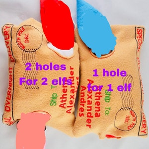 May include: Two tan felt elf pouches with red and blue elf hats. The pouches have text that reads "Overnight Delivery" and "North Pole Dec." One pouch has "2 holes for 2 elves" and the other has "1 hole for 1 elf."