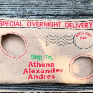May include: A beige felt envelope with red and black embroidered details. The text "SPECIAL OVERNIGHT DELIVERY" is at the top. It has two circular cutouts and the text "Ship To: Athena Alexander Andres" in red and green.