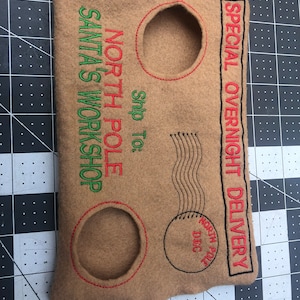 May include: Tan felt envelope with embroidered text and details. Features the phrases "North Pole," "Santa's Workshop," and "Special Overnight Delivery." Includes two circular cutouts and a postage stamp design. Red and green thread accents.