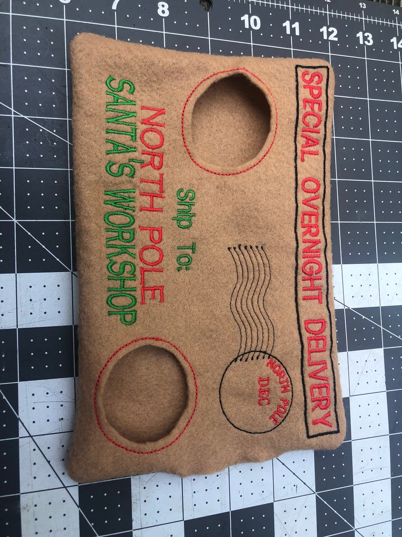 May include: Tan felt envelope with embroidered text and details. Features the phrases "North Pole," "Santa's Workshop," and "Special Overnight Delivery." Includes two circular cutouts and a postage stamp design. Red and green thread accents.