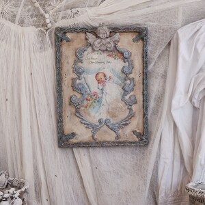 May include: A vintage-style display featuring a framed artwork with a baby illustration, a decorative urn, a cross, and a small vase. The color palette includes soft whites, creams, and pinks, with lace and beaded accents. The overall theme is a delicate, antique aesthetic.