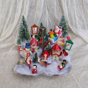 May include: A vintage Christmas diorama with miniature figures of Santa, reindeer, children, and snowmen. The scene includes small houses, trees, and street lamps, set against faux snow. The colour scheme is dominated by red, green, and white, evoking a festive atmosphere.