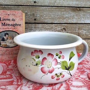 Op de afbeelding: Witte emaille kamerpot met een handvat, versierd met roze en rode bloemmotieven en groene bladeren. Een vintage Frans kookboek getiteld "Le Livre de la Ménagère" is zichtbaar op de achtergrond. De pot staat op een rood-wit patroon doek.