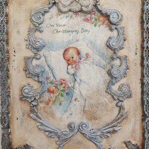May include: A vintage-style baptismal plaque with a distressed finish. The plaque features a central image of a baby surrounded by floral accents and the text "On Your Christening Day." The frame includes an angel and ornate scrollwork.