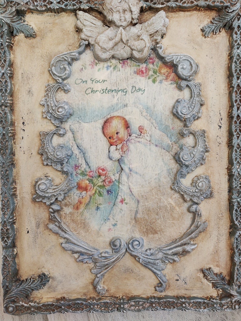 May include: A vintage-style baptismal plaque with a distressed finish. The plaque features a central image of a baby surrounded by floral accents and the text "On Your Christening Day." The frame includes an angel and ornate scrollwork.