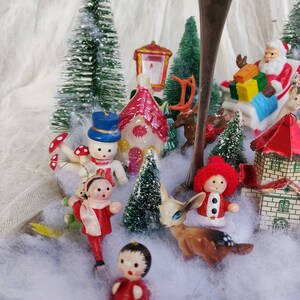 May include: A Christmas-themed display with miniature figurines including Santa Claus, a snowman, houses, and deer. The scene is set amongst small, snow-covered trees and artificial snow, creating a charming festive display.
