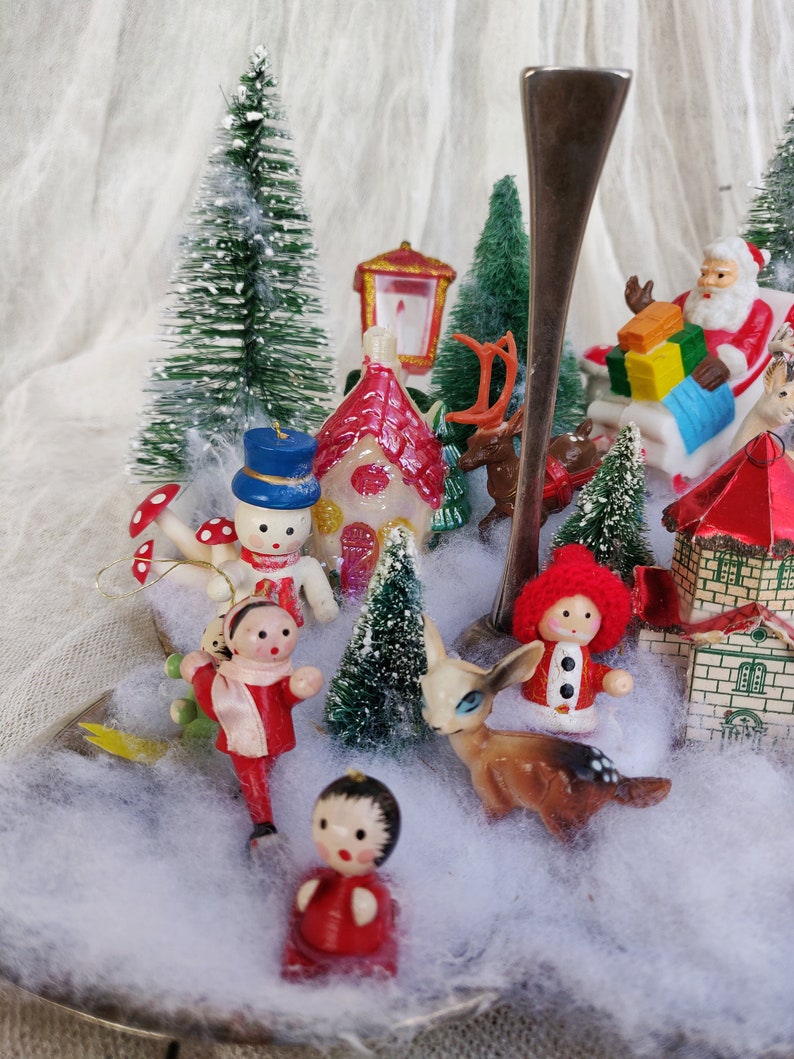 May include: A Christmas-themed display with miniature figurines including Santa Claus, a snowman, houses, and deer. The scene is set amongst small, snow-covered trees and artificial snow, creating a charming festive display.