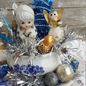 May include: A vintage Christmas centerpiece featuring a blue tinsel tree, porcelain angel figurines, and silver tinsel. The arrangement is set in a white and blue floral teacup and saucer, with decorative ornaments.