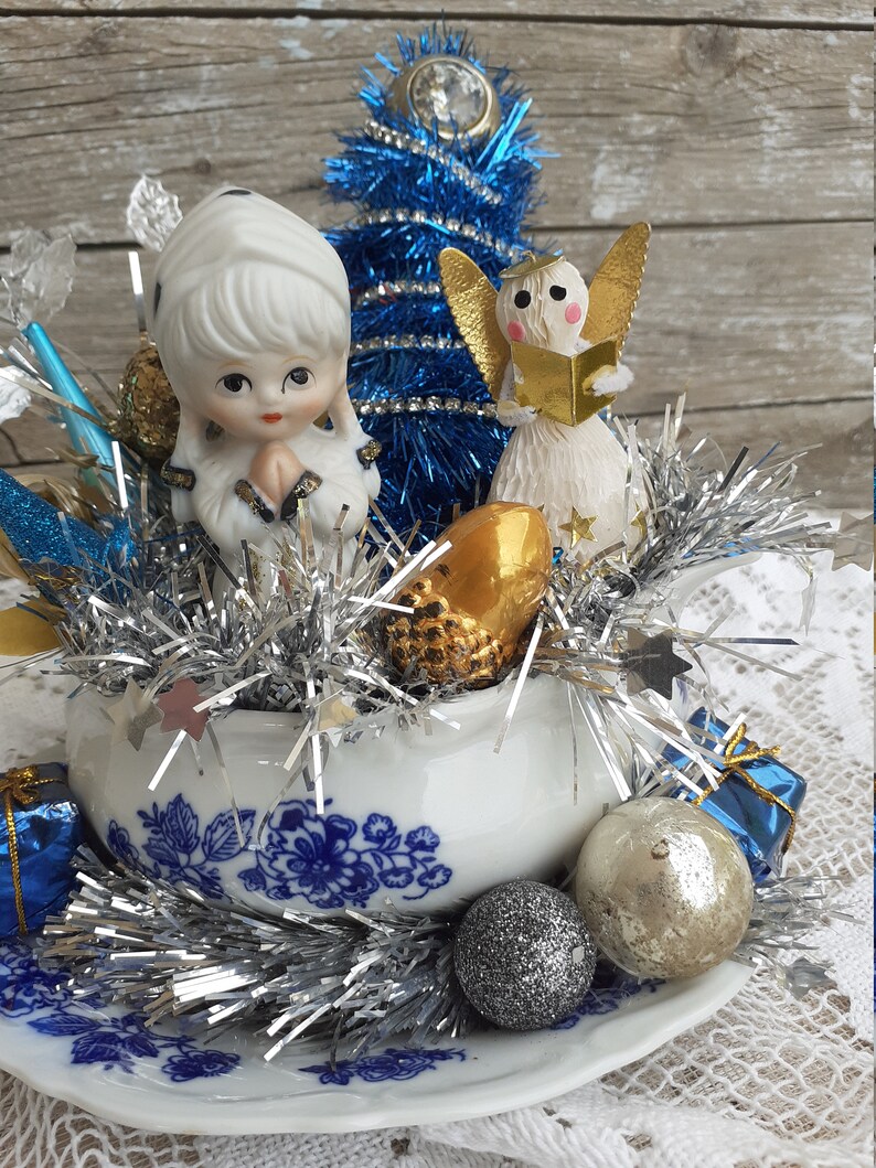 May include: A vintage Christmas centerpiece featuring a blue tinsel tree, porcelain angel figurines, and silver tinsel. The arrangement is set in a white and blue floral teacup and saucer, with decorative ornaments.