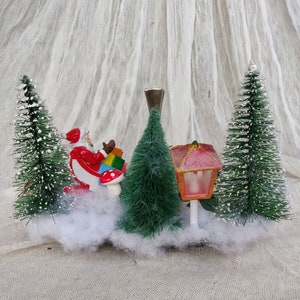 May include: A festive Christmas miniature scene. It features Santa Claus with gifts, bottle brush trees, and a red and pink lantern. The scene is set on a bed of white fluff, creating a holiday display.