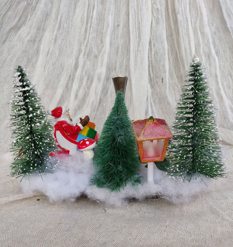 May include: A festive Christmas miniature scene. It features Santa Claus with gifts, bottle brush trees, and a red and pink lantern. The scene is set on a bed of white fluff, creating a holiday display.