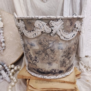 Può includere: Un secchio decorativo in stile vintage con finitura anticata. Il secchio presenta finiture e manici bianchi e ornati. Il corpo del secchio è dipinto con un disegno di cherubini sbiadito. Il secchio è appoggiato su una pila di vecchi libri.