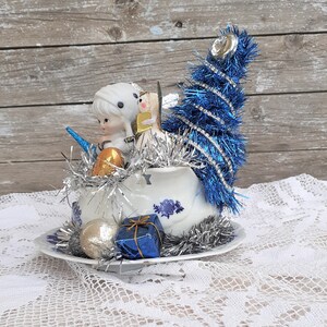 May include: A vintage Christmas decoration featuring a white teacup with blue floral designs, filled with silver tinsel, small figurines, and a blue tinsel Christmas tree. Additional ornaments include a gold and silver ball, and a blue gift box.