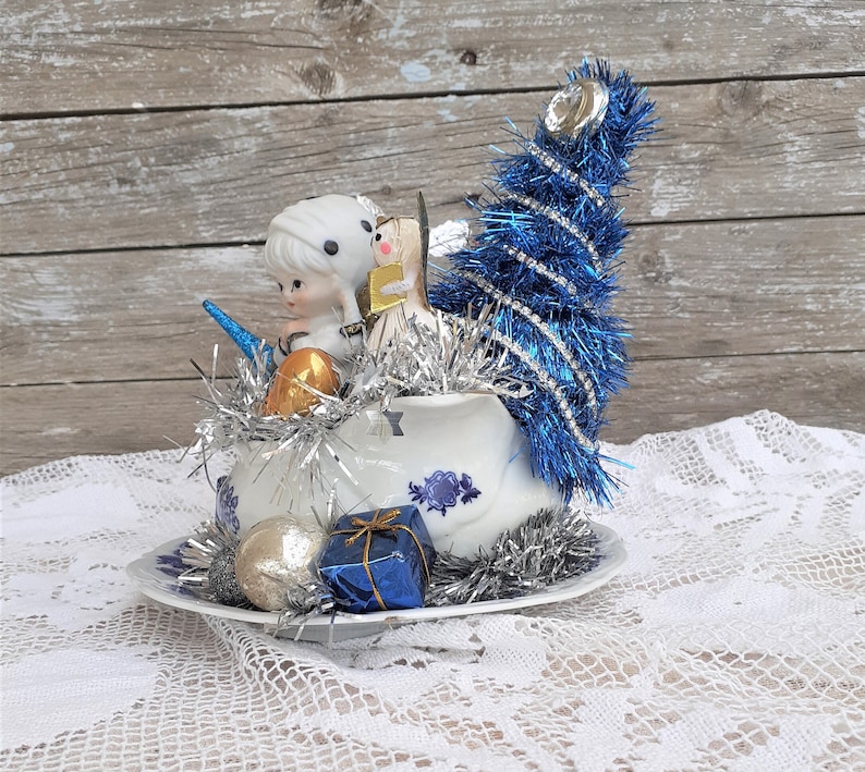 May include: A vintage Christmas decoration featuring a white teacup with blue floral designs, filled with silver tinsel, small figurines, and a blue tinsel Christmas tree. Additional ornaments include a gold and silver ball, and a blue gift box.