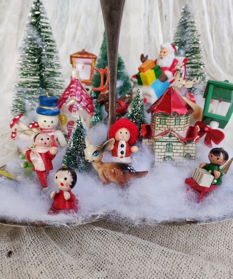 May include: A Christmas-themed display on a silver serving tray, featuring miniature figurines and bottle brush trees. The scene includes Santa Claus, reindeer, houses, snowmen, and other festive ornaments, creating a charming winter wonderland.