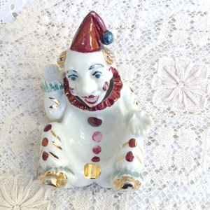 May include: A white ceramic clown figurine with a red and gold hat, red polka dots, and gold accents. The clown has a smiling face with blue eyes and a red ruffled collar. The figurine is in a seated position.