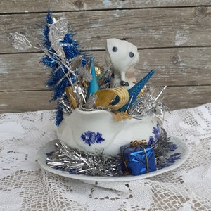 May include: A vintage Christmas decoration featuring a white porcelain shoe filled with silver and blue tinsel, ornaments, and a small figurine. A blue gift box sits on the saucer, which has blue floral designs. The background is a wooden surface.
