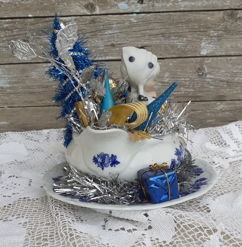 May include: A vintage Christmas decoration featuring a white porcelain shoe filled with silver and blue tinsel, ornaments, and a small figurine. A blue gift box sits on the saucer, which has blue floral designs. The background is a wooden surface.
