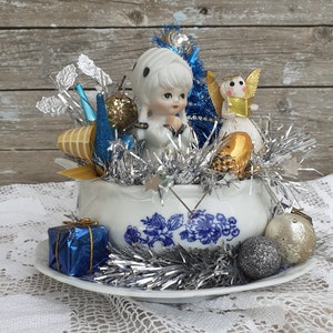 May include: A vintage Christmas centerpiece featuring a porcelain doll, angel figurines, and ornaments in a blue and white floral bowl. Silver tinsel, blue and gold accents, and a wrapped gift box complete the festive arrangement.
