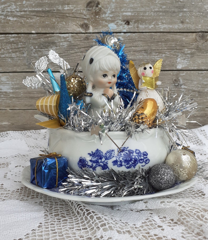May include: A vintage Christmas centerpiece featuring a porcelain doll, angel figurines, and ornaments in a blue and white floral bowl. Silver tinsel, blue and gold accents, and a wrapped gift box complete the festive arrangement.