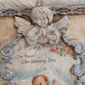May include: A vintage-style baptism plaque featuring a praying angel figurine at the top. The plaque has a decorative frame and includes the text "On Your Christening Day" above an illustration of a baby.