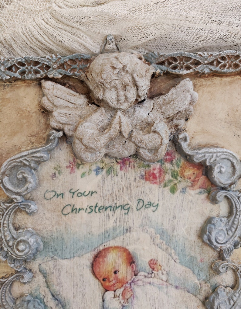 May include: A vintage-style baptism plaque featuring a praying angel figurine at the top. The plaque has a decorative frame and includes the text "On Your Christening Day" above an illustration of a baby.