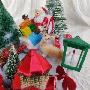 May include: A festive miniature Christmas scene with Santa Claus in a sleigh, a reindeer, and a small house with a red roof. The scene includes miniature green trees, a green lantern, and a red bow. The scene is set on a white surface.