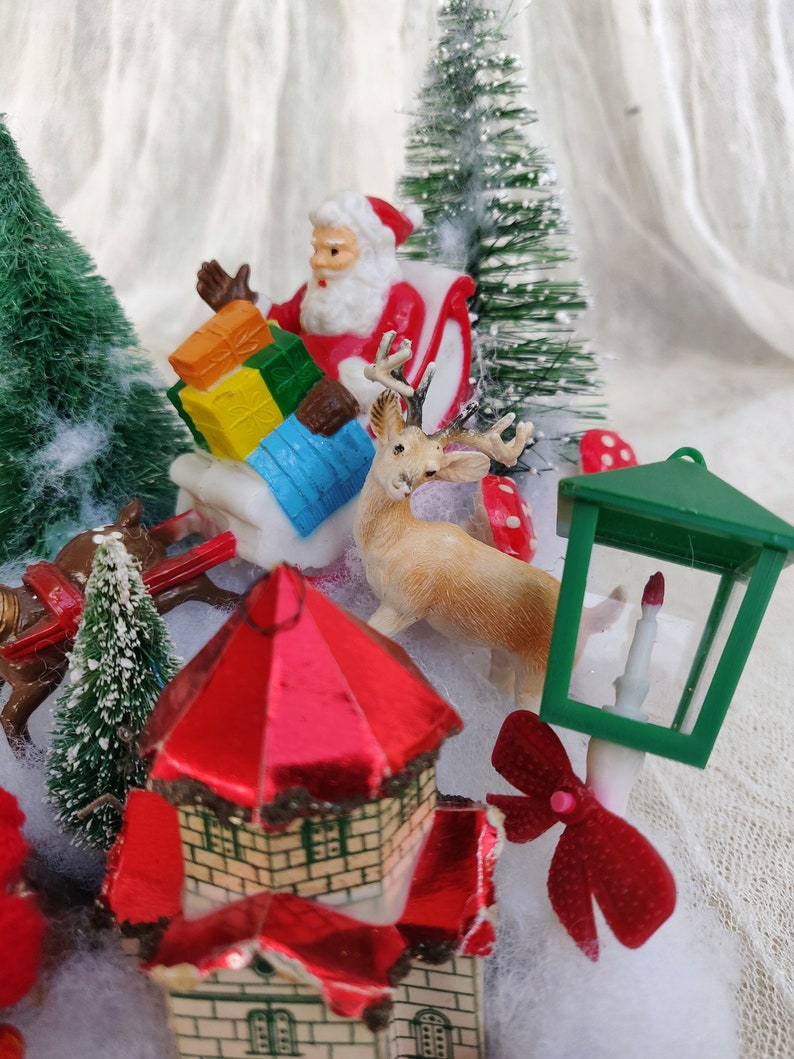 May include: A festive miniature Christmas scene with Santa Claus in a sleigh, a reindeer, and a small house with a red roof. The scene includes miniature green trees, a green lantern, and a red bow. The scene is set on a white surface.