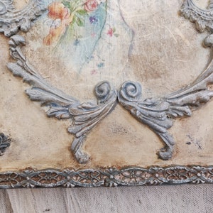May include: A decorative wooden plaque with a vintage aesthetic. The plaque features a faded floral print in pastel colors, framed by ornate, silver-colored relief detailing. The edges have a distressed, aged appearance, adding to the antique charm.