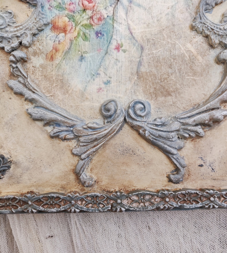 May include: A decorative wooden plaque with a vintage aesthetic. The plaque features a faded floral print in pastel colors, framed by ornate, silver-colored relief detailing. The edges have a distressed, aged appearance, adding to the antique charm.