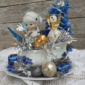 May include: A vintage Christmas centerpiece featuring a white and blue floral patterned creamer and saucer. The arrangement includes a porcelain doll, a paper angel, blue tinsel, silver garland, gold acorn, and blue gift boxes. Silver and gold ornaments complete the festive display.