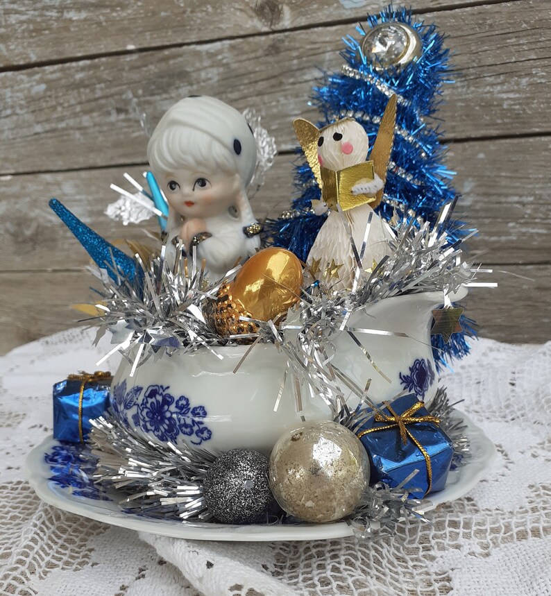 May include: A vintage Christmas centerpiece featuring a white and blue floral patterned creamer and saucer. The arrangement includes a porcelain doll, a paper angel, blue tinsel, silver garland, gold acorn, and blue gift boxes. Silver and gold ornaments complete the festive display.