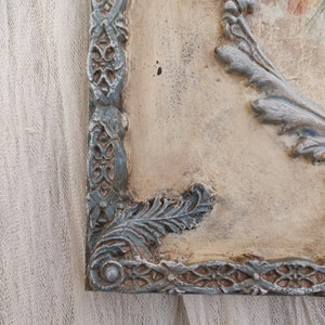 May include: A corner view of a decorative wall plaque with a distressed finish. The plaque features a light beige background with ornate, raised details in shades of gray and teal. The frame has a detailed, floral-inspired design.