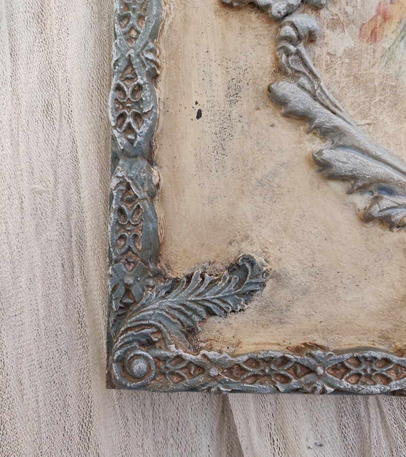 May include: A corner view of a decorative wall plaque with a distressed finish. The plaque features a light beige background with ornate, raised details in shades of gray and teal. The frame has a detailed, floral-inspired design.
