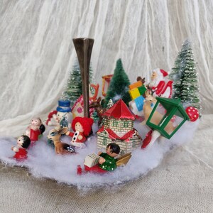 May include: A festive Christmas display featuring miniature figurines of Santa, a snowman, and children, surrounding a central candle holder. The scene includes small houses, trees, and a lantern, set on a bed of artificial snow.